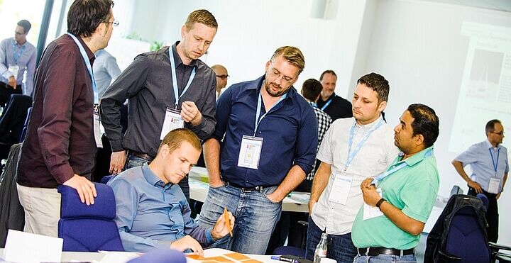 eine Gruppe von Teilnehmenden am Prof. Scrum Master Kurs-1 (PSM1) die gemeinsam ein Daily Scrum Meeting durchführen. Einer der Personen sitzt am Tisch andere stehen drum herum