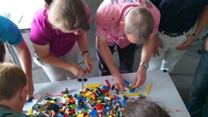 viele Teilnehmer in einem Scrum Workshop für alle Rollen stehen um einen Tisch und simulieren mit Lego einen Sprint