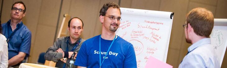 Jan Fischbach (Trainer und Berater bei Scrum-Events) moderiert einen Open-Space Workshop. Teilnehmer stehen vor einem Flipp Chart und diskutieren.
